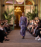 Model walking down a runway in an elegant indoor setting with chandeliers and spectators.