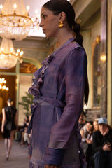 Woman in a purple outfit standing in an elegant indoor setting with chandeliers.