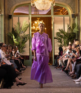 Model in a purple outfit walking on a runway with chandeliers and spectators in the background.