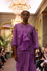 Model wearing a purple outfit with ruffled details in an elegant indoor setting.