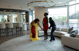 Three women interacting in a modern living room with a chandelier and large windows.