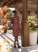 Woman in a pink dress standing under a wooden pergola with plants and chairs in the background.