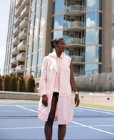 Woman in a light pink coat standing on a tennis court with tall buildings in the background