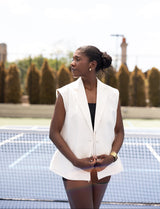 Woman in a white vest standing on a tennis court with trees in the background