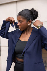 Woman wearing a navy blue blazer over a black top, standing against a light-colored wall.
