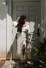Woman in a white dress standing in front of a white door with sunlight casting shadows.