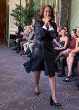 Woman in a black dress with sheer sleeves walking in an indoor setting with people seated in the background.